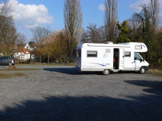 Wohnmobilstellplatz am Klunsberg Nieheim Weißes Wohnmobil auf einem ruhigen Parkplatz, umgeben von Bäumen und blauen Himmel mit Wolken.