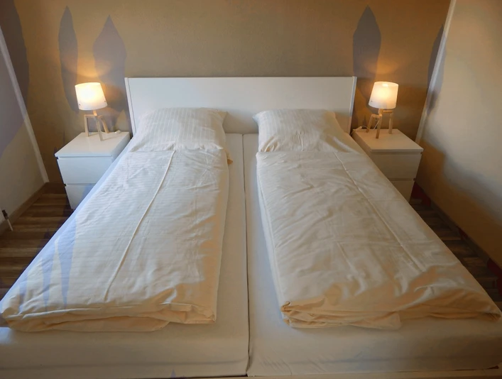 Ferienwohnung SchiederSee Hell eingerichtetes Doppelbett mit weißen Bettlaken, flankiert von zwei Nachttischen mit Lampen.