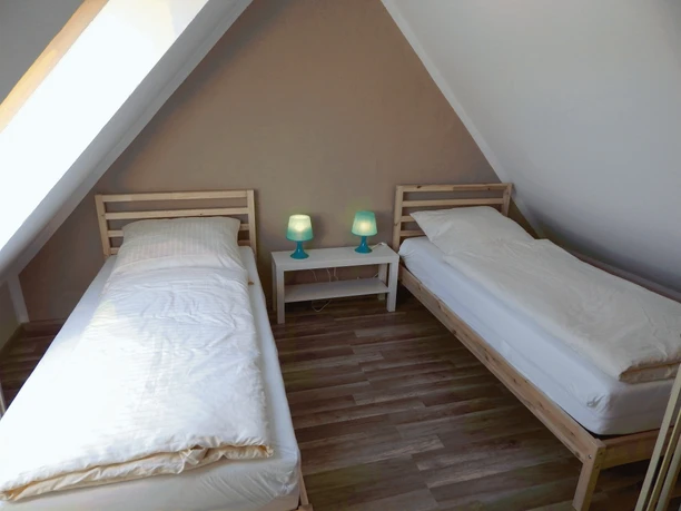 Zwei Einzelbetten in einem Dachzimmer mit schräge Wände, dazwischen ein kleiner Tisch mit Lampen.