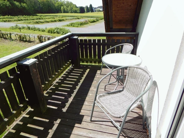 Zwei Rattan-Stühle und ein runder Tisch auf einem Holzbalkon mit Blick in eine grüne Landschaft.