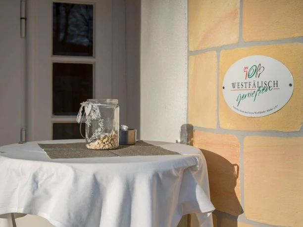 Westfälisch genießen im Landgasthof Potthoff, Borgholzhausen-Barnhausen Ein liebevoll dekorierter Stehtisch mit einem Glasgefäß, daneben eine Plakette mit der Aufschrift "Westfälisch genießen".