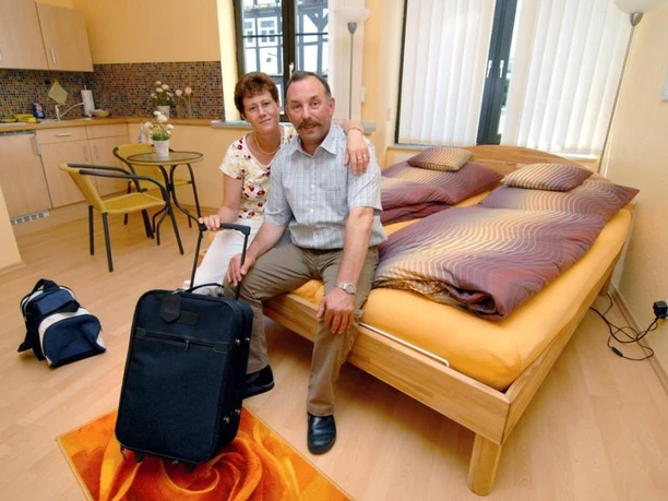 Ankommendes Ehepaar, sitzend auf hochwertigem Bett, Im Hintergrund Küchenzeile und Bistrotisch mit Stühlen