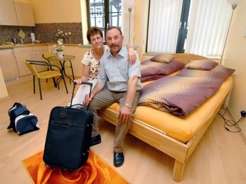 Zimmer Ankommendes Ehepaar, sitzend auf hochwertigem Bett, Im Hintergrund Küchenzeile und Bistrotisch mit Stühlen