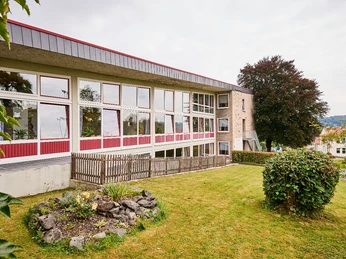 Moderne Jugendherberge mit großflächigen Fenstern, umgeben von einer gepflegten Rasenfläche und Bäumen.