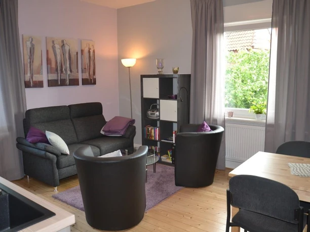 Modernes Wohnzimmer mit grauem Sofa, zwei schwarzen Sesseln, Bücherregal und großem Fenster.