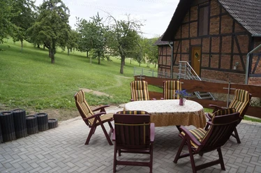 Grillplatz/ Außenterasse Sechs gemütliche Holzstühle umrahmen einen runden Tisch auf einer gepflasterten Terrasse vor einer Wiese.