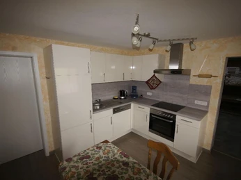 Moderne Küche mit weißen Hochglanz-Schränken, Elektroherd und Esstisch mit blumiger Tischdecke.