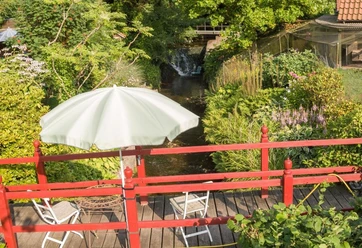 Brücke zum Garten über den Ilsenbach, im Hintergrund der Wasserfall Rot gestrichene Holzbrücke über den Ilsenbach, umgeben von üppigem Garten, dahinter ein Wasserfall.