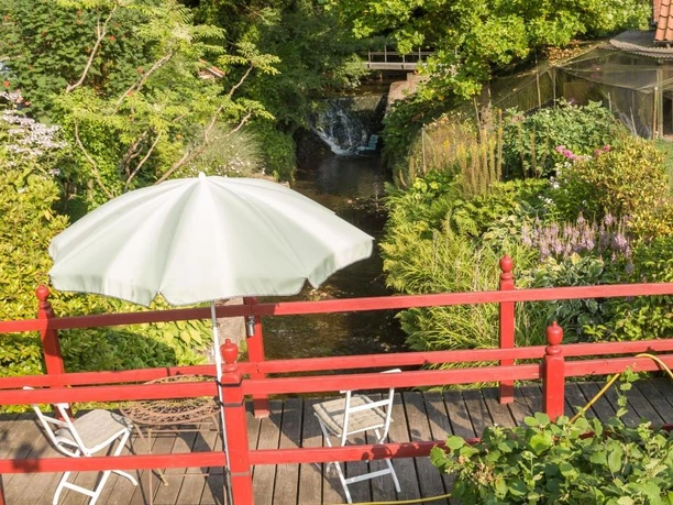 Brücke zum Garten über den Ilsenbach, im Hintergrund der Wasserfall Rot gestrichene Holzbrücke über den Ilsenbach, umgeben von üppigem Garten, dahinter ein Wasserfall.