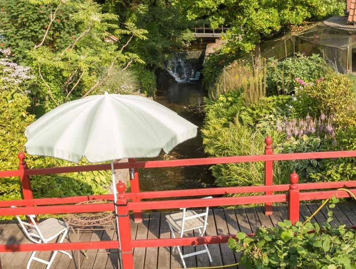 Brücke zum Garten über den Ilsenbach, im Hintergrund der Wasserfall Rot gestrichene Holzbrücke über den Ilsenbach, umgeben von üppigem Garten, dahinter ein Wasserfall.