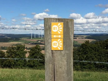 Wegweiserpfosten zeigt Wanderroute "Blickwinkel" in einer weitläufigen Landschaft mit Windrädern.
