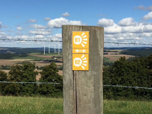 Wegweiserpfosten zeigt Wanderroute "Blickwinkel" in einer weitläufigen Landschaft mit Windrädern.
