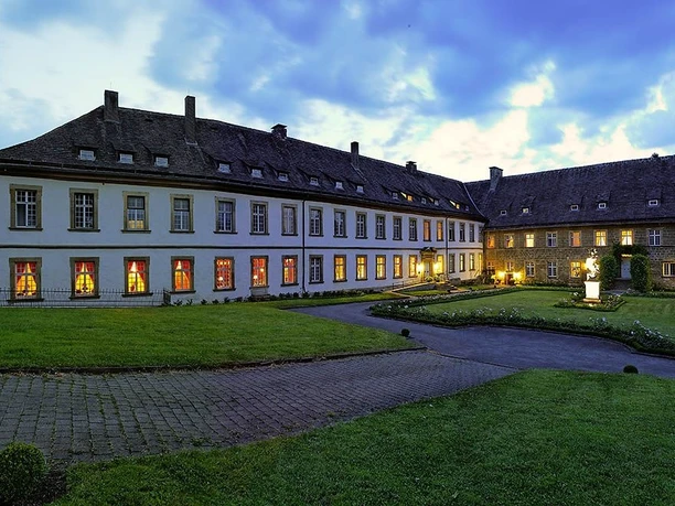 Beleuchtetes Schloss Gehrden bei Dämmerung, umgeben von gepflegtem Rasen und wolkigem Himmel.