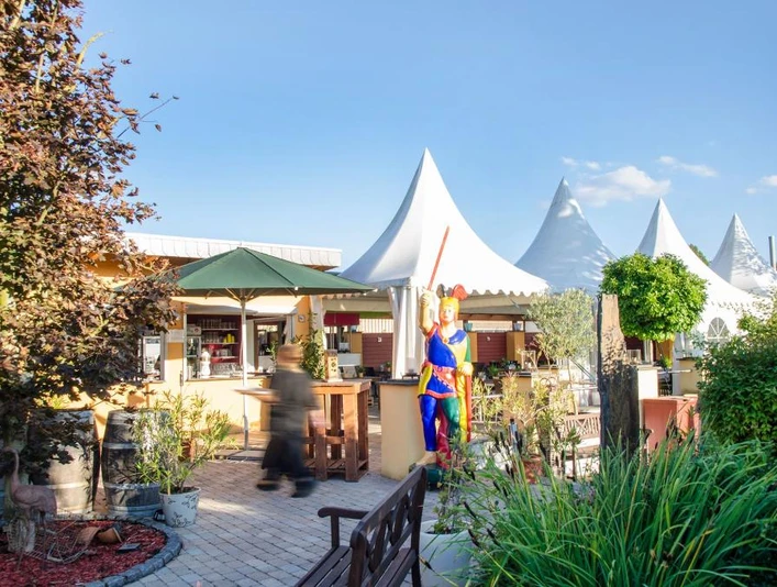 Biergarten mit Sonnenschirmen und Sitzplätzen, geschmückt mit Pflanzen, Skulptur in bunter Kleidung.