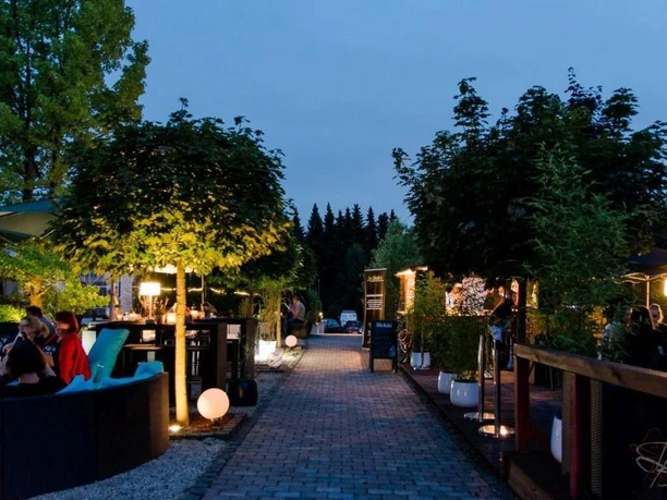 Biergarten Ein beleuchteter Biergarten bei Abenddämmerung mit Gästen auf gepflastertem Weg und umgeben von Bäumen.