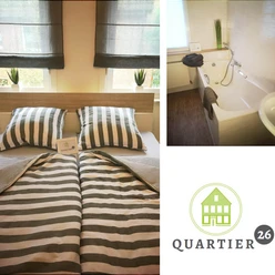 Doppelbett mit gestreifter Bettwäsche in hellem Raum, daneben Badezimmer mit moderner Badewanne.