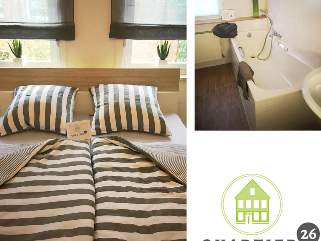 fewo-quartier26 Doppelbett mit gestreifter Bettwäsche in hellem Raum, daneben Badezimmer mit moderner Badewanne.