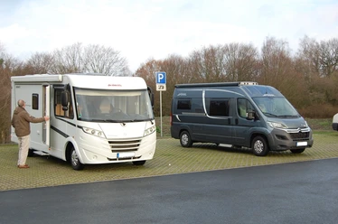 Zwei Wohnmobile parken auf einem gepflasterten Parkplatz vor kahlen Bäumen unter bewölktem Himmel.