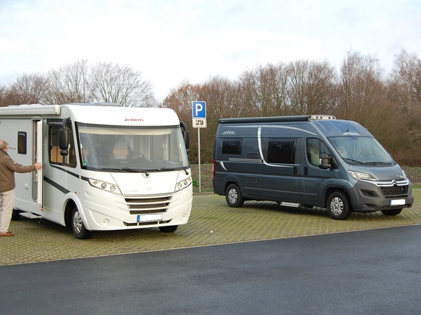 Wohnmobilstellplätze in Herzebrock-Clarholz Zwei Wohnmobile parken auf einem gepflasterten Parkplatz vor kahlen Bäumen unter bewölktem Himmel.