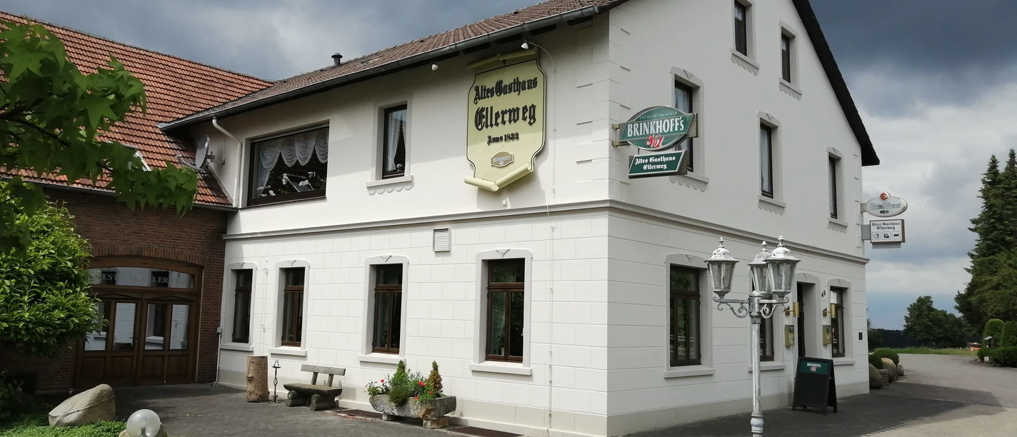 altes_gasthaus_ellerweg_teutoburger-wald-tourismus_bohlken Historisches zweistöckiges Gasthaus mit weißer Fassade, umgeben von gepflastertem Hof und grüner Landschaft.