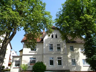 Das zweistöckige Haus mit mansardartigem Dach liegt zwischen grünen Bäumen, umgeben von blauen Himmel.