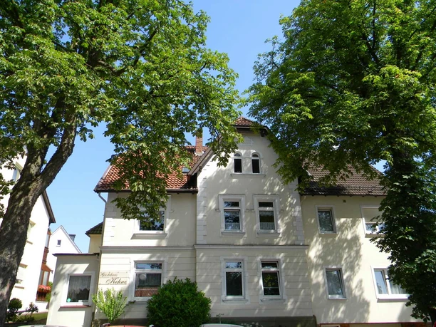 Das zweistöckige Haus mit mansardartigem Dach liegt zwischen grünen Bäumen, umgeben von blauen Himmel.
