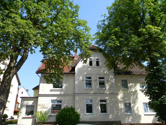 Das zweistöckige Haus mit mansardartigem Dach liegt zwischen grünen Bäumen, umgeben von blauen Himmel.