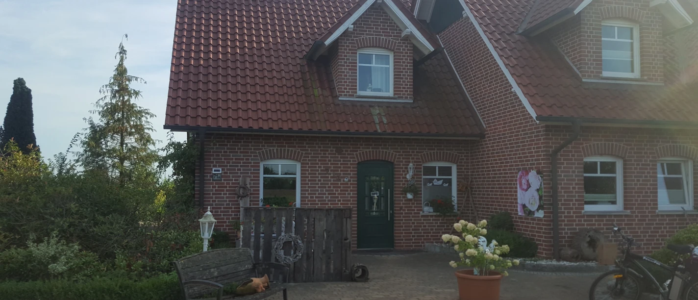 ferienwohnung-emsland-20190823_175728_001 Backsteinhaus mit rotem Ziegeldach, grünem Eingang und bepflanzter Einfahrt im Abendlicht.