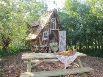 Kleines, schiefes Holzhaus inmitten grüner Bäume; Vordergrund: Picknicktisch mit Speisen.