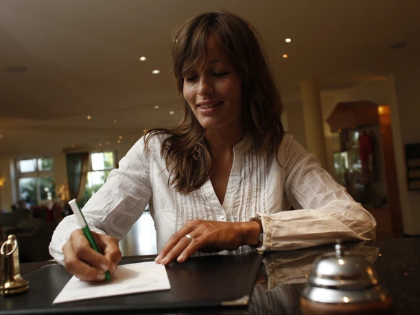 Eine Frau schreibt in einer Hotelrezeption lächelnd auf Papier, umgeben von eleganter Einrichtung.