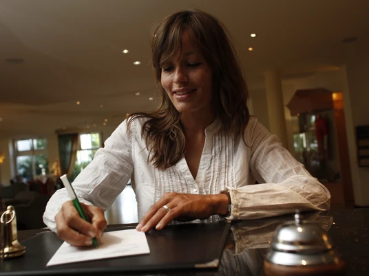 platzhalterbild_uebernachtung_twt_t-conrad Eine Frau schreibt in einer Hotelrezeption lächelnd auf Papier, umgeben von eleganter Einrichtung.