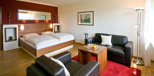 hotel_rosenberg_bad-driburg_1600 Modernes Hotelzimmer mit Doppelbett, Ledersofas, Couchtisch, Sektkühler, hellem Holzboden und Wandkunst.