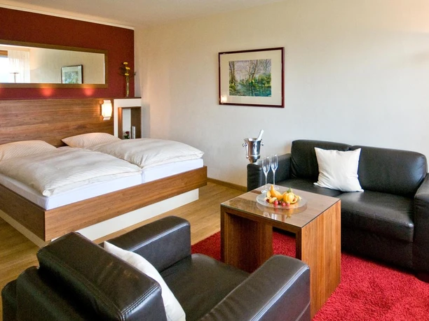 hotel_rosenberg_bad-driburg_1600 Modernes Hotelzimmer mit Doppelbett, Ledersofas, Couchtisch, Sektkühler, hellem Holzboden und Wandkunst.