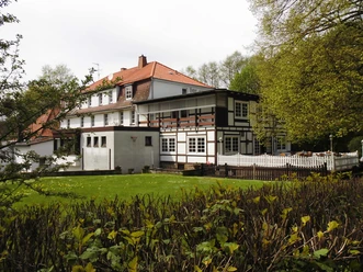 aussenansicht_haus-varlemann_warburg Fachwerkhaus mit rotem Ziegeldach, umgeben von Bäumen und Hecken, mit gepflegtem Rasen im Vordergrund.
