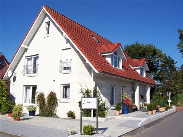 pension-haus-emsblick-haren Zweistöckiges Haus mit rotem Satteldach, hellen Wänden und bepflanztem Eingangsbereich bei Sonne.