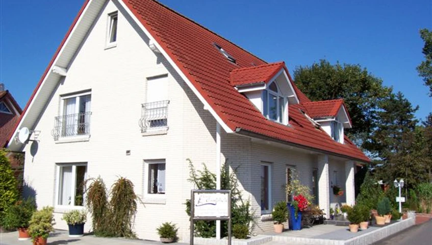 pension-haus-emsblick-haren Zweistöckiges Haus mit rotem Satteldach, hellen Wänden und bepflanztem Eingangsbereich bei Sonne.