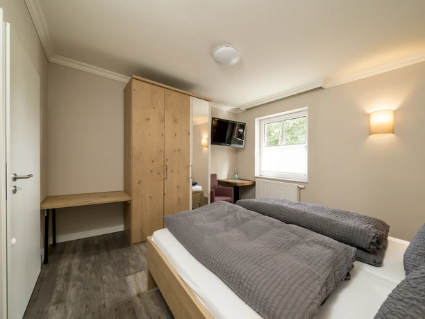 2019_07_10_dsc3061 Modern eingerichtetes Doppelzimmer mit Holzmöbeln, grauer Bettwäsche und Blick auf Fenster und Fernseher.