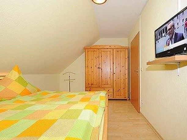 haren-schlazi-c_nst_6808_kl Helles Schlafzimmer mit Doppelbett, Holzmöbeln und Wandfernseher unter der Dachschräge.