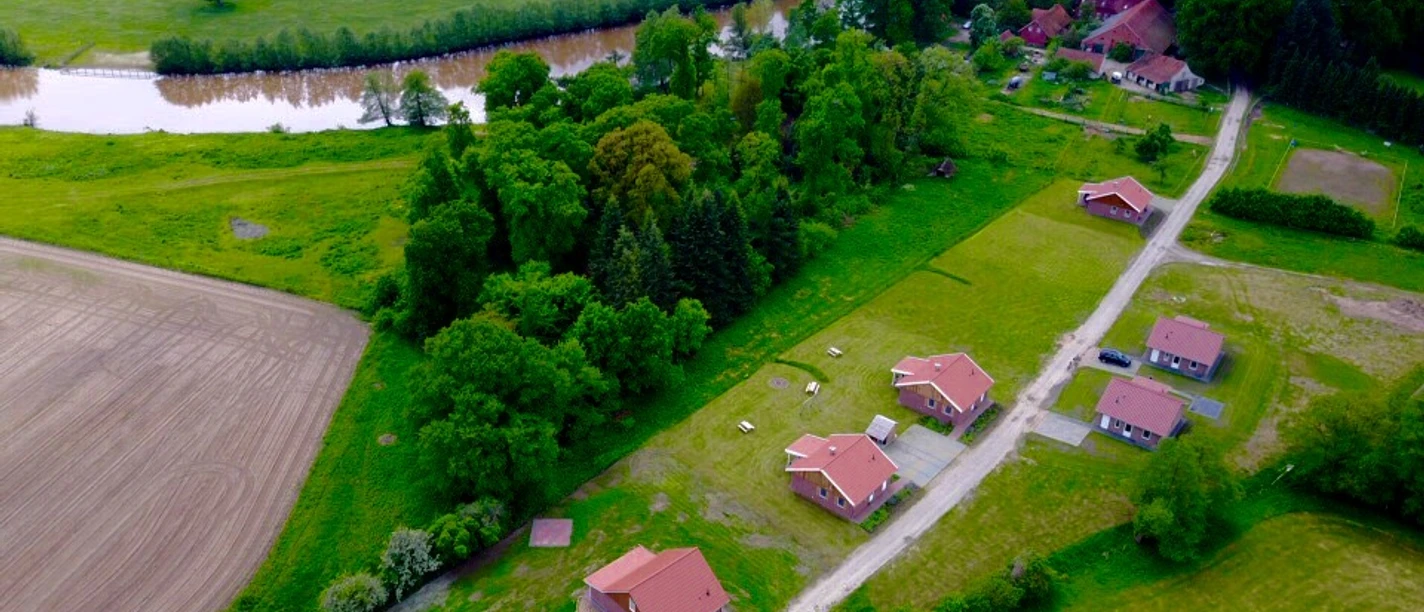 fereinh-user-emsland-luftbild-1 Luftaufnahme mehrerer Ferienhäuser mit roten Dächern, umgeben von Wiesen, Feldern und Flusslandschaft.