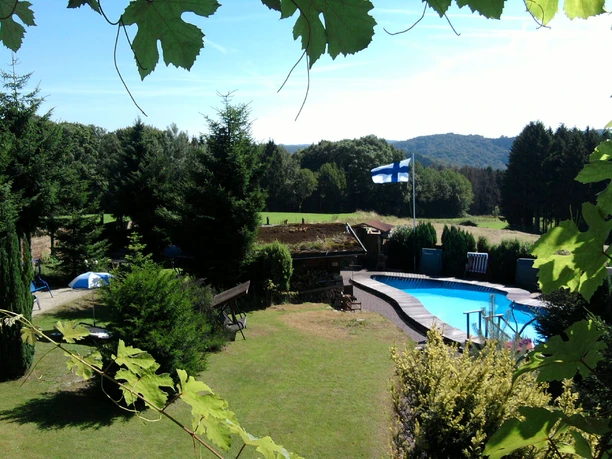 Eifgen-Sauna Ein malerischer Garten mit Pool, umgeben von Bäumen, im Hintergrund sanfte Hügel unter blauem Himmel.
