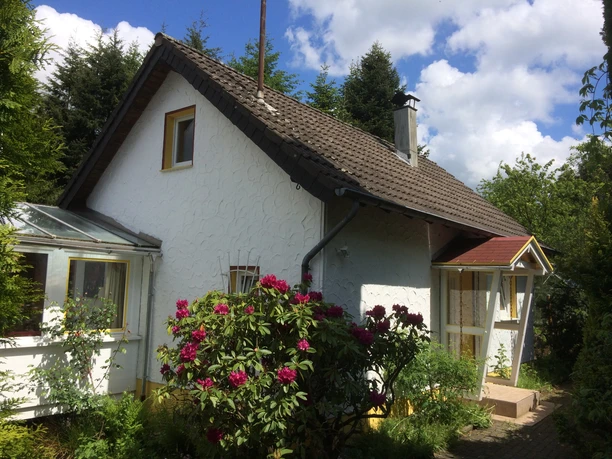 Ferienhaus Ketzberg Ein weißes Einfamilienhaus mit Satteldach, umgeben von blühenden Rhododendron und grünen Bäumen.