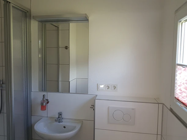 Büschem's Box Barrierefreies Badezimmer mit Waschbecken, Spiegel, Duschkabine und Fenster mit Faltrollo.