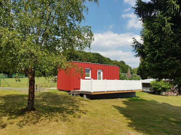 Büschem's Box Rotes Ferienhaus mit Terrasse, umgeben von Wald und Wiese an einem sonnigen Tag. Entspannende Idylle.