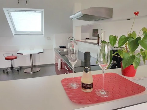 Ferienwohnung "Bergisches Land" Moderne Küchenküche mit weißer Arbeitsplatte, Champagnerflasche, zwei Gläsern, Tisch und Pflanze.