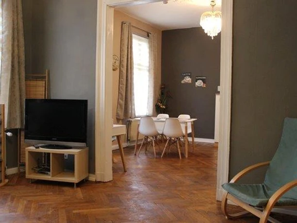 Blick zum Essbereich Wohnzimmer mit Holzboden, Fernseher, Schaukelstuhl und Essbereich mit weißem Tisch und Stühlen.