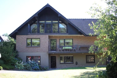 Ferienwohnung "Zum Nutscheid" Zweistöckiges, rotes Ziegelhaus mit Balkon und Garten, umgeben von grüner Vegetation unter blauem Himmel.