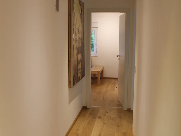 Blick auf Küche Ein schmaler Flur mit Holzboden und weiß gestrichenen Wänden, führt zu einem Zimmer mit Tageslicht.