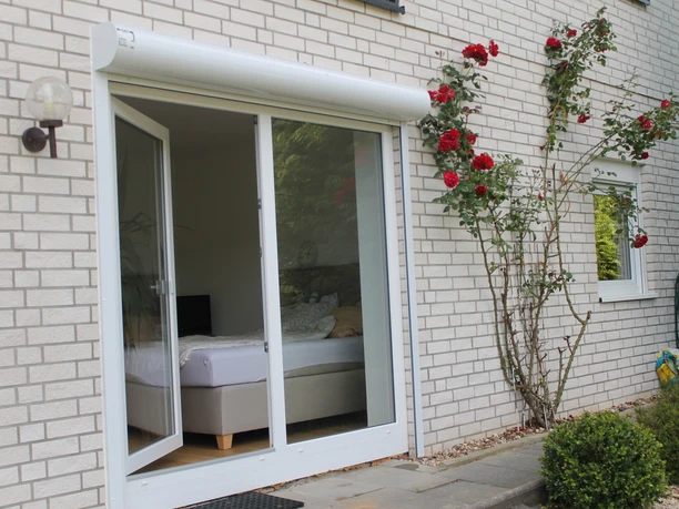 Außenansicht Terrassentür eines Hauses geöffnet, zeigt gemütliches Schlafzimmer im Inneren, Kletterrosen an der Fassade.