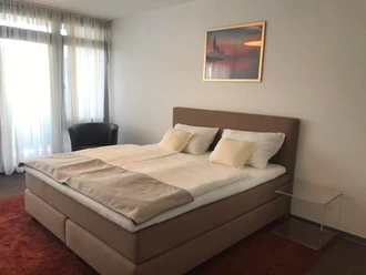 Modernes Hotelzimmer mit Doppelbett, weißen und beigen Bettdecken, samt schwarzem Sessel und Glastisch.