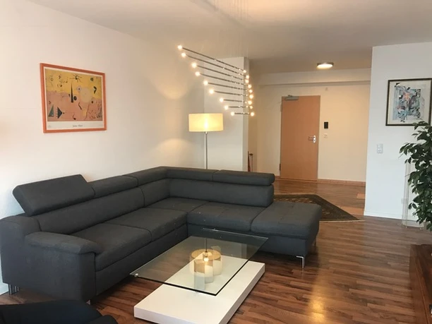 Ferienwohnung bei Prinz Modernes Wohnzimmer mit grauer Eckcouch, Glastisch, Bodenlampe und stilvollen Kunstwerken an der Wand.